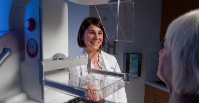 A female Allied Health staff member stands beside a mammogram machine, talking with a female patient.