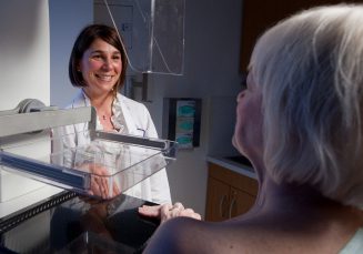 A female Allied Health staff member stands beside a mammogram machine, talking with a female patient.