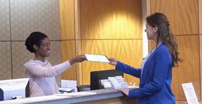 Two female administrative staff members work and talk with each other in a hospital reception setting.