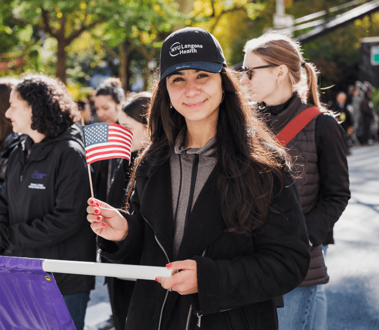 A young female staff member stands and smiles in an outdoor setting as she waves a small U.S. flag.