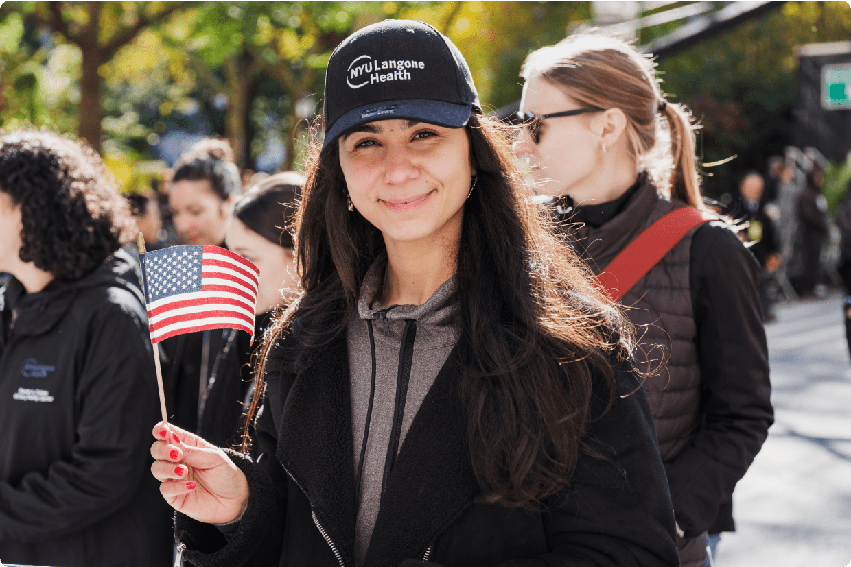 A young female staff member stands and smiles in an outdoor setting as she waves a small U.S. flag.