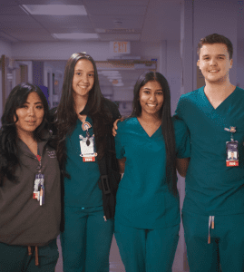 Four young male and female NYU Langone nursing staff stand together smiling in a hospital ward setting.