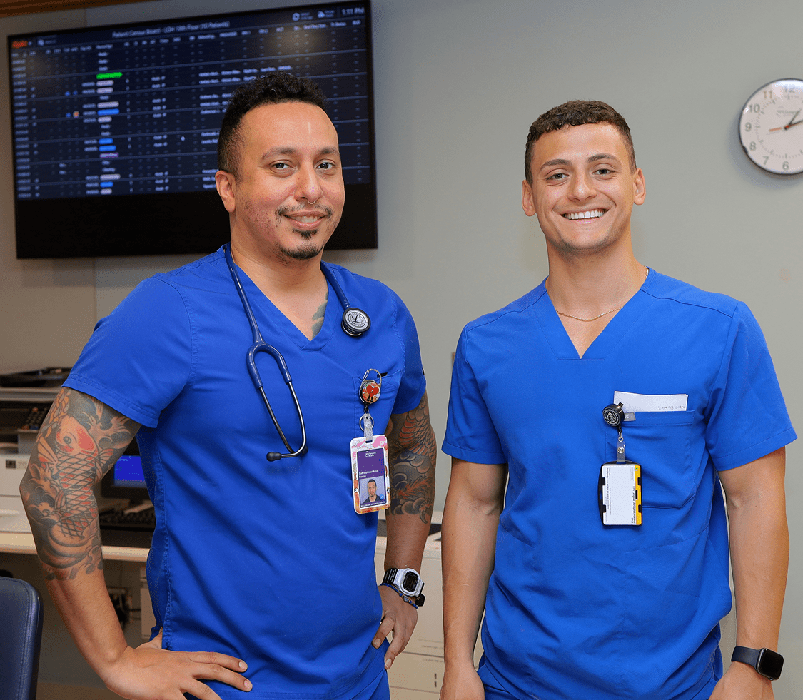 Two male NYU Langone nursing staff members stand together smiling in a hospital ward setting.