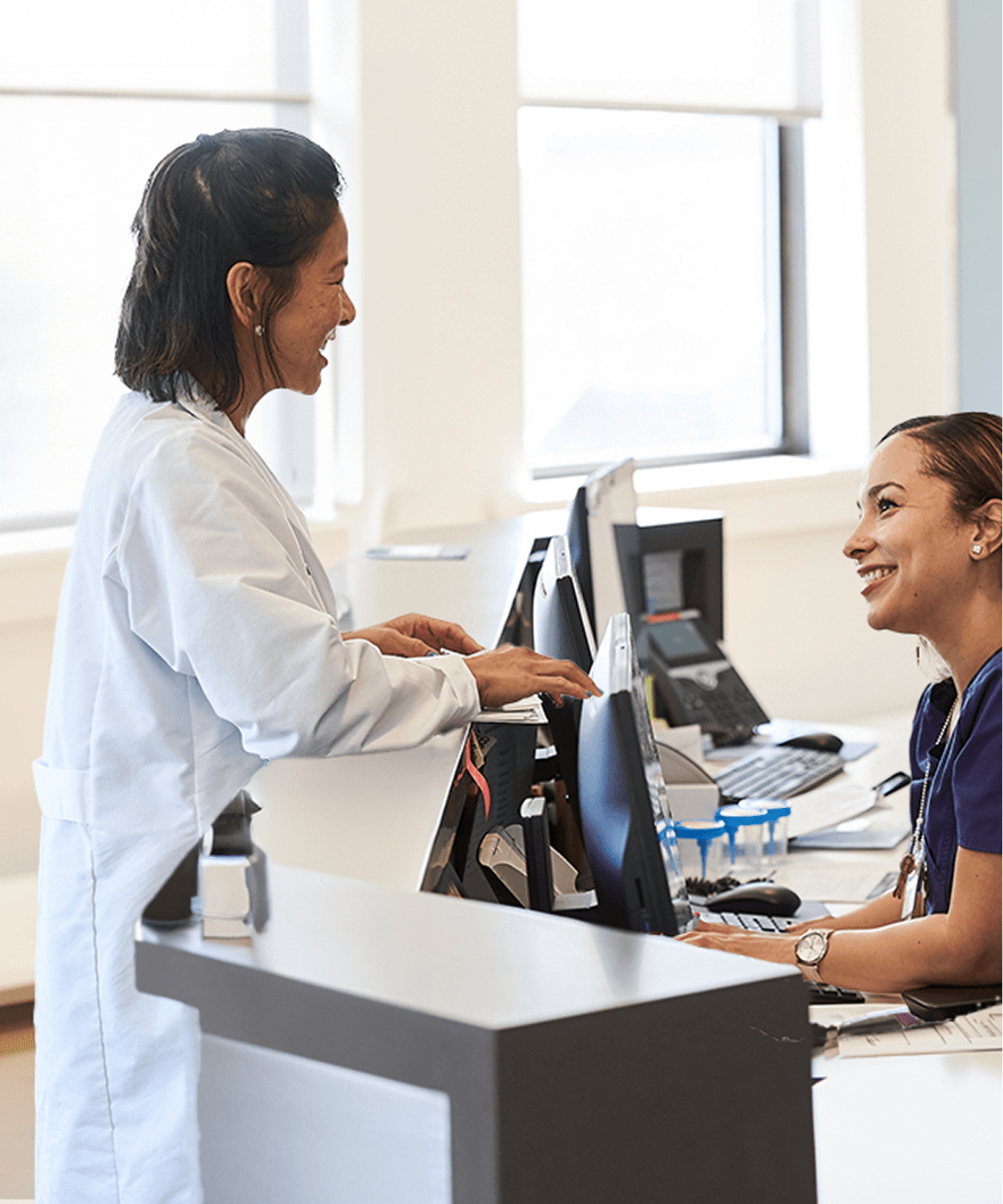 Two female NYU Langone staff members smile and talk together at a hospital reception desk.