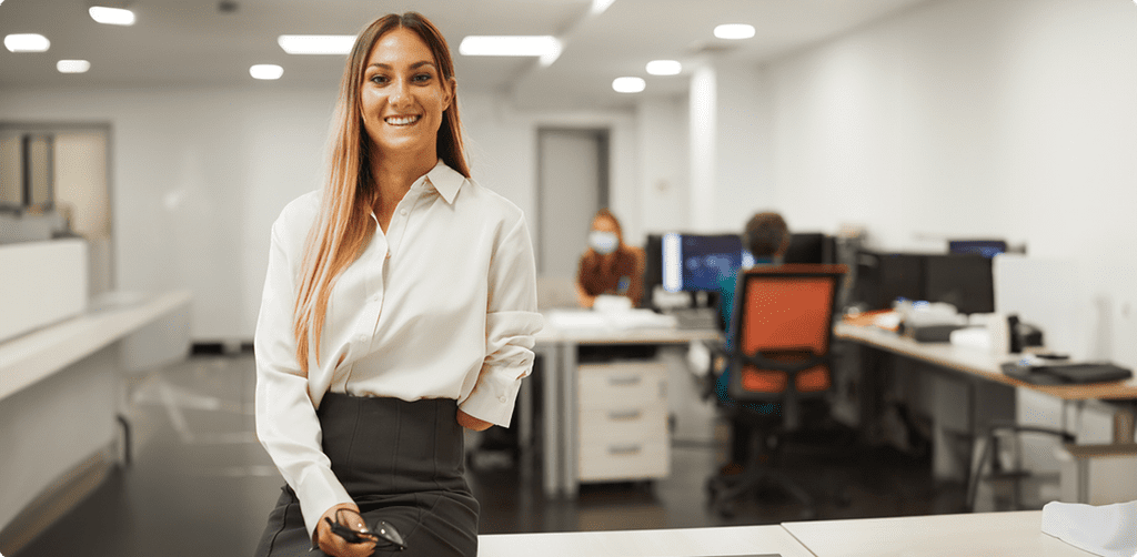 A smiling woman with a left-arm limb difference standing in a modern office with colleagues working in the background.