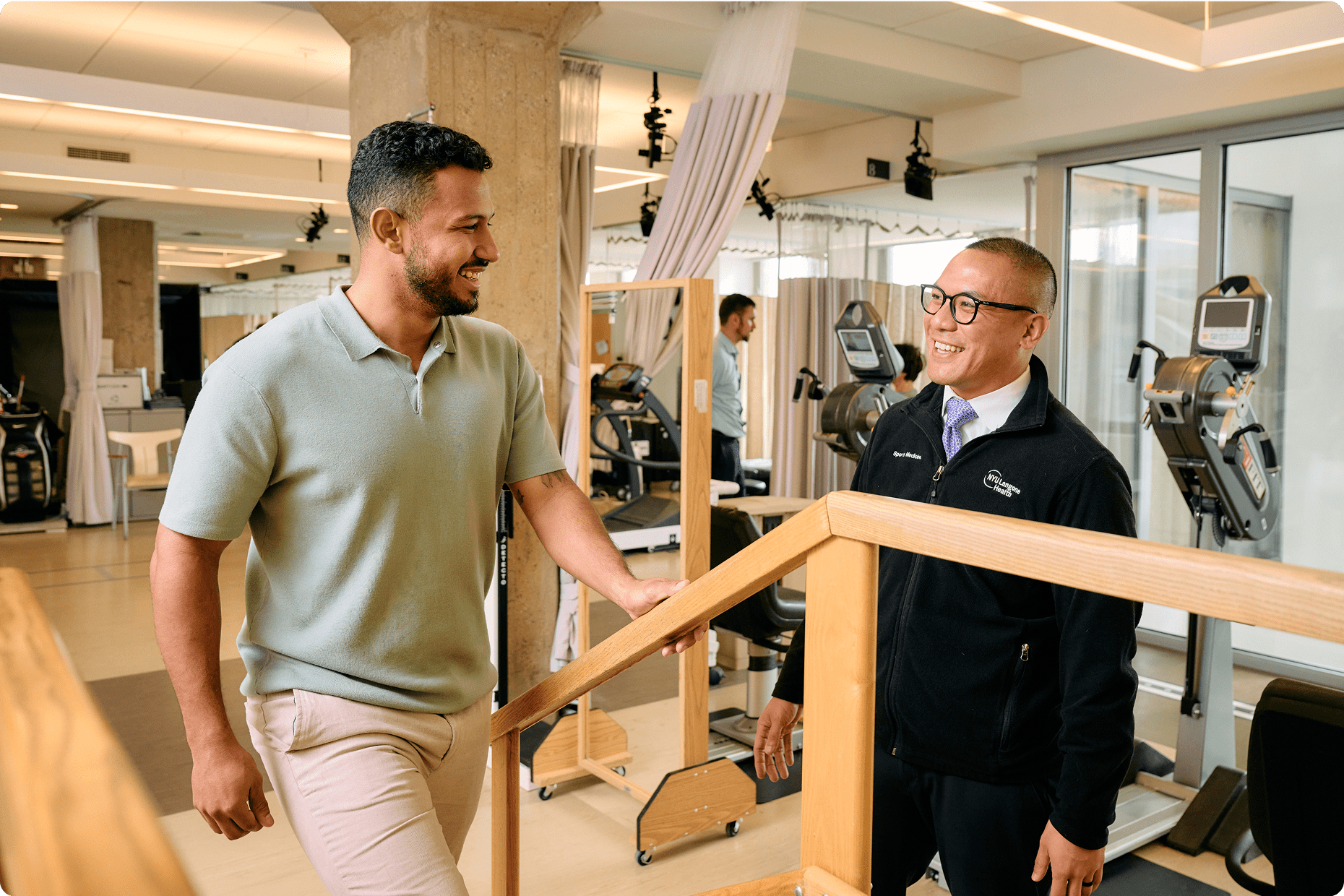A male patient climbs stairs in a rehab therapy room while an NYU Langone Orthopedic Hospital staff member encourages him.