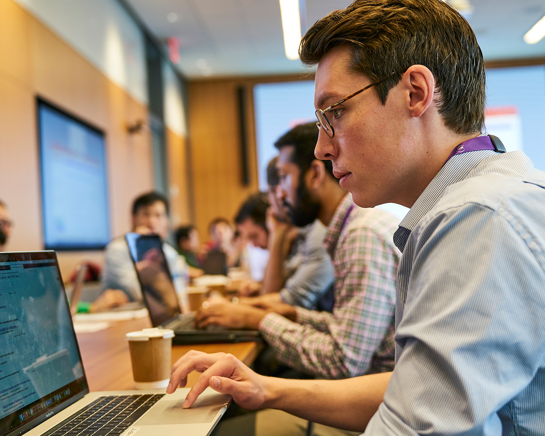 A male staff member at NYU Langone looks at a computer screen while sitting in a room with other computer-focused staff.