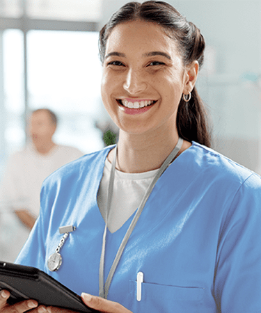 A smiling healthcare professional wearing light blue scrubs stands in the foreground with a doctor and patient having a conversation in the background.