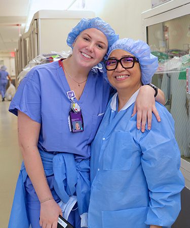 Two nurses from the NYU Langone Orthopedic Hospital nursing unit wearing light blue colored scrubs embrace one another.